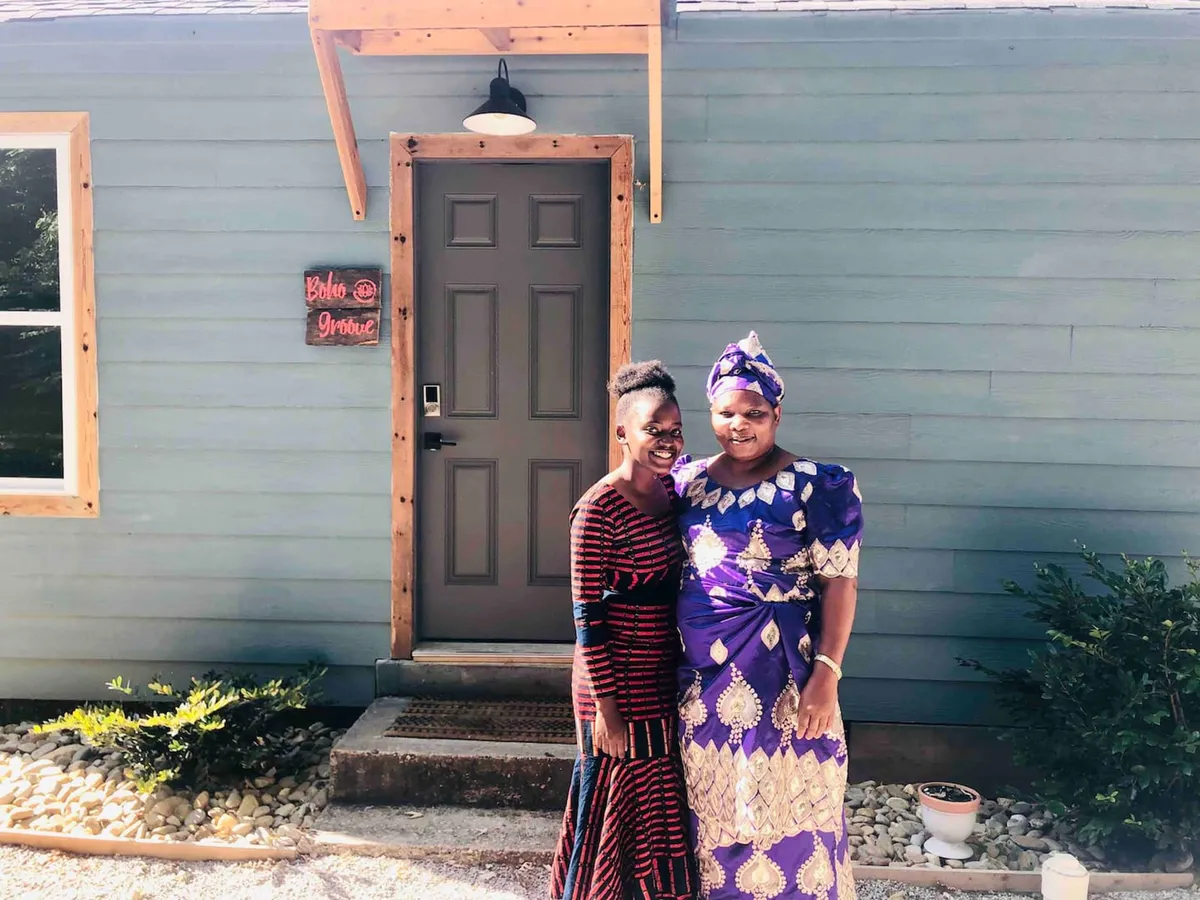 The image features the front door and a sign on the wall, with two women in traditional attire greeting guests. The joyful energy and focus on the entrance highlight the warmth and attentiveness of the hosts.