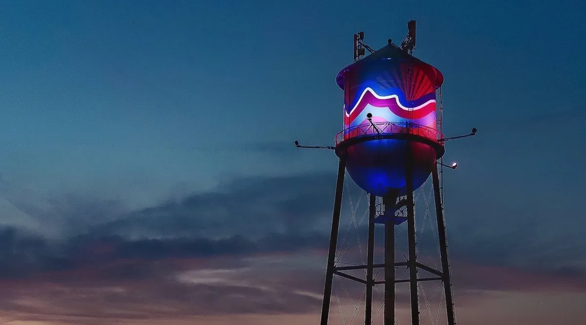 A whimsical twilight view of the neighborhood’s iconic water tower, glowing with colored lights against a dusky sky. The area below hints at community gatherings and evening activities, inviting visitors for strolls and photography.