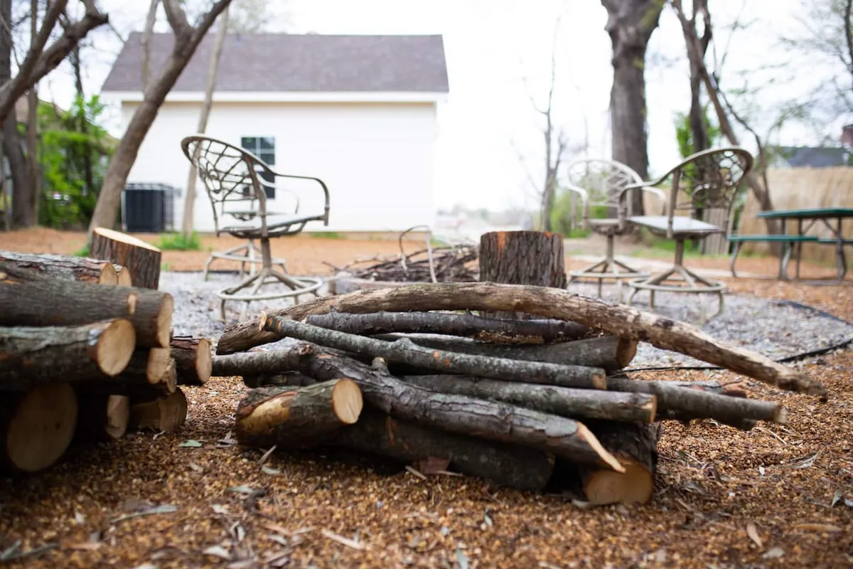 A cozy backyard scene featuring wooden logs arranged around a fire pit, with garden chairs and a gravel path cutting through lush greenery. The setting is welcoming and shaded, evoking the sense of a hidden oasis perfect for gatherings or quiet reflection.