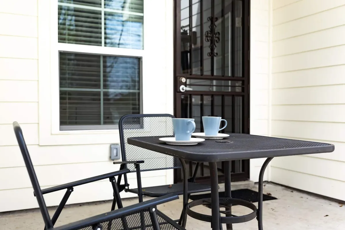 This image showcases an outdoor table and chairs set on a patio, with coffee cups set out, suggesting a tranquil spot for morning relaxation or casual conversation. The scene is bright, calm, and surrounded by greenery, capturing the essence of a peaceful backyard retreat.