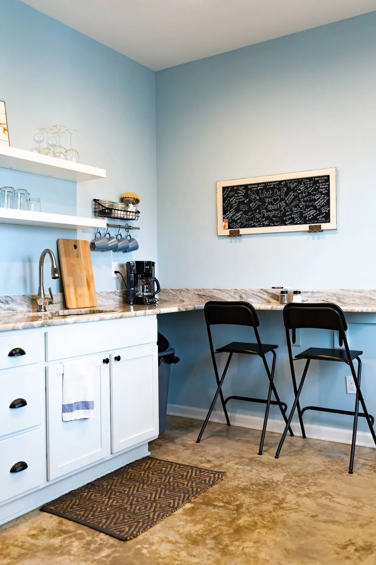 A cozy kitchenette with a broad countertop, a chalkboard for notes or local recommendations, a coffee maker, and bar stools. The scene exudes casual comfort, inviting guests to enjoy coffee, meals, or conversation in a relaxed setting.