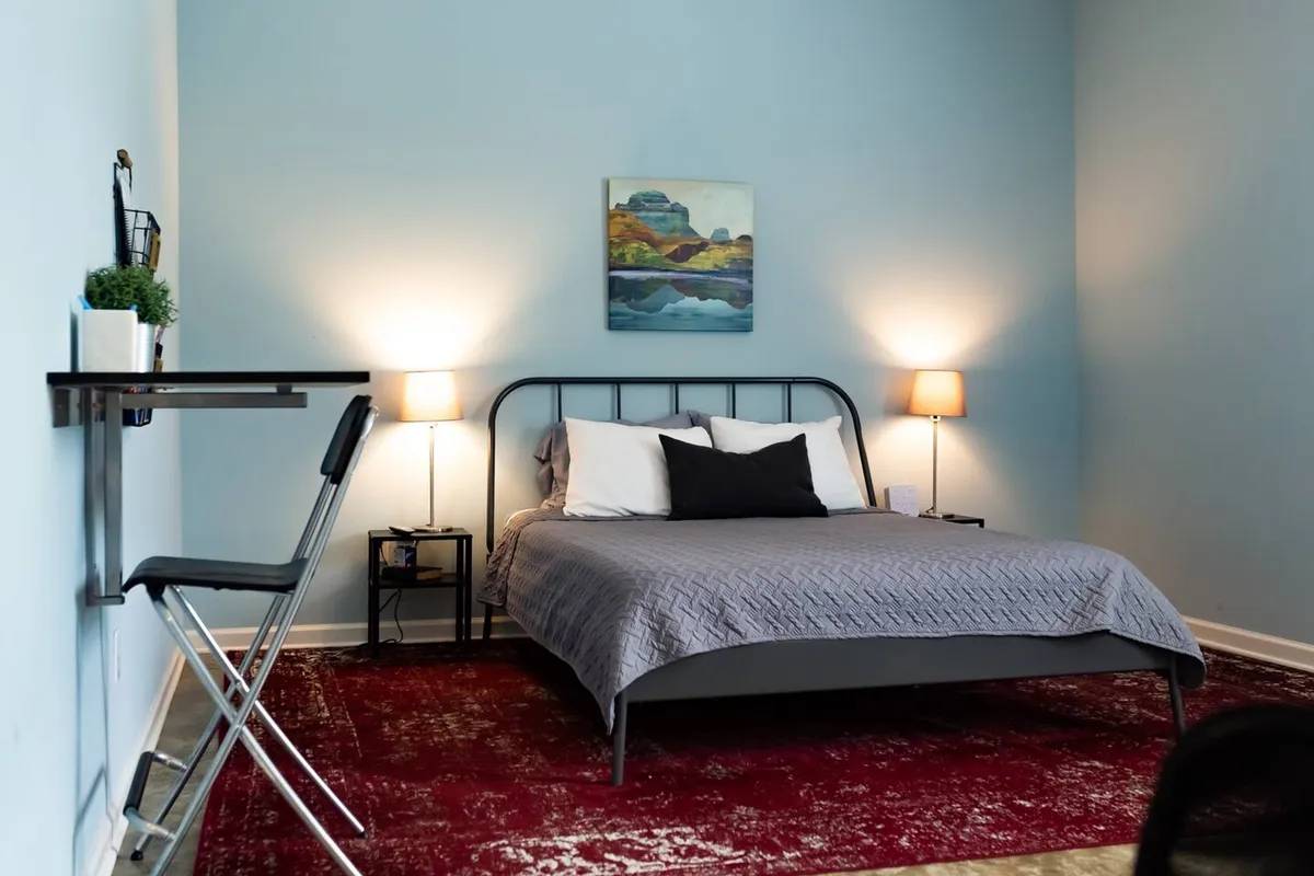 A tranquil bedroom scene with a plush bed as the focal point, accented by tasteful artwork above. A table and chair provide a spot for reading or remote work, while soft lighting from lamps creates an intimate, soothing atmosphere.