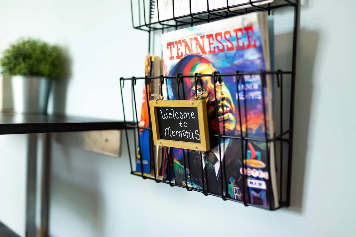 A bright corner of the studio featuring a 'Welcome to Memphis' sign, a magazine rack stocked with reading materials, and a lush green plant. The arrangement suggests both a sense of arrival and connections to the local community, making the space feel more inviting for newcomers.
