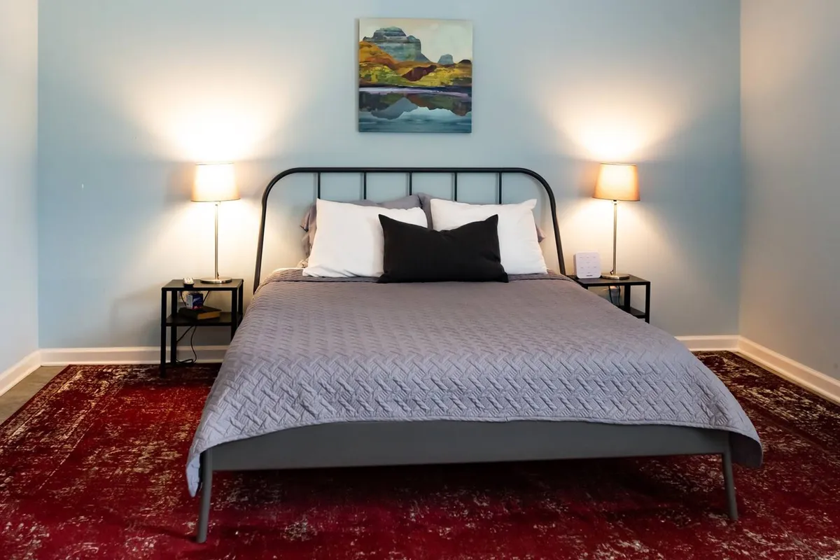 A cozy bedroom featuring a plush bed with soft linens, flanked by nightstands and warm lamp light. Artwork adds a creative touch above the headboard, and a large rug lends comfort to the space. Natural light gently filters in, creating a serene atmosphere perfect for relaxation.