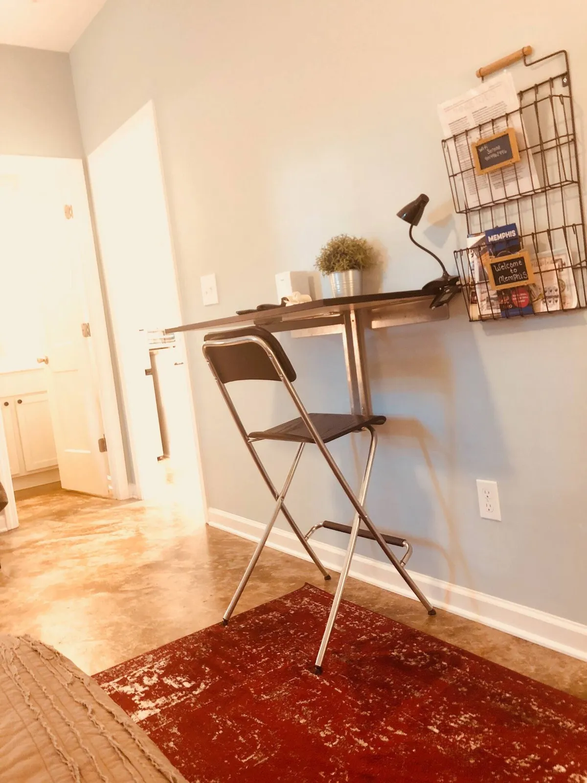 A bright interior work area featuring a folding desk paired with a black chair, illuminated by a modern metal lamp. A wire wall organizer holds notes and accessories, and a small potted plant adds a touch of greenery. The clean setup suggests an ideal space for productivity and focus.