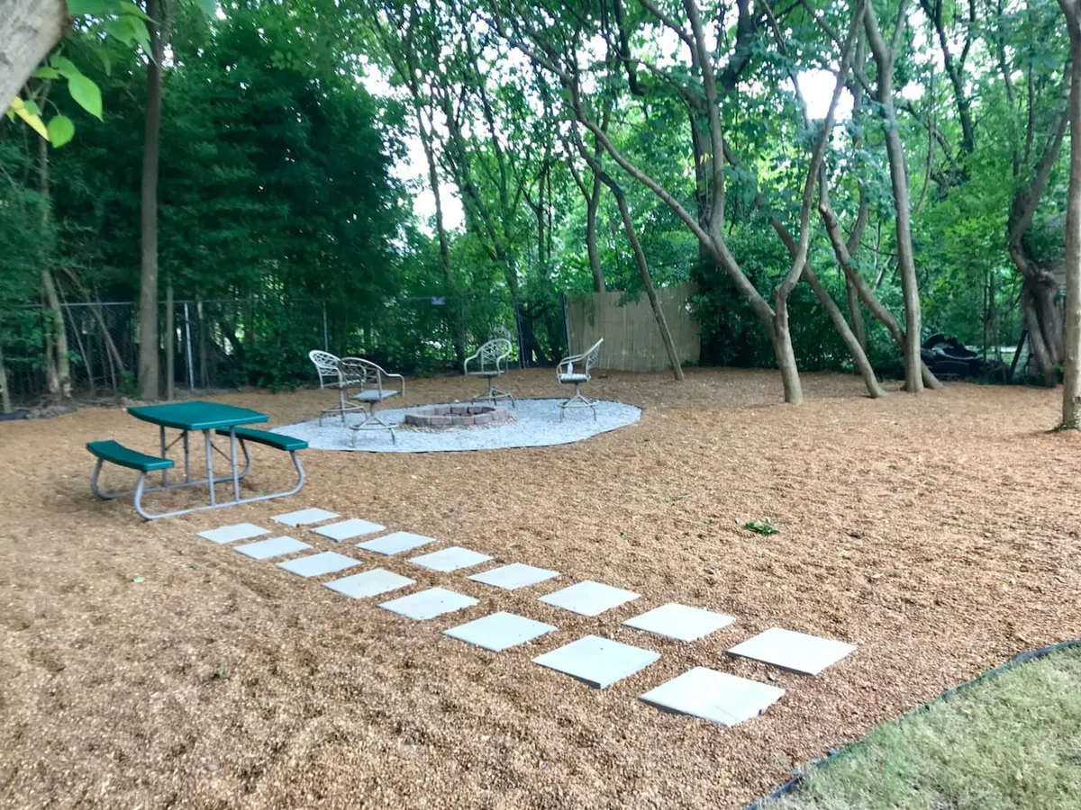 A relaxed backyard scene featuring a fire pit surrounded by inviting seating and a winding pathway, with greenery and dappled shade creating a tranquil outdoor environment. The space exudes comfort and is perfect for gatherings or quiet moments outdoors.