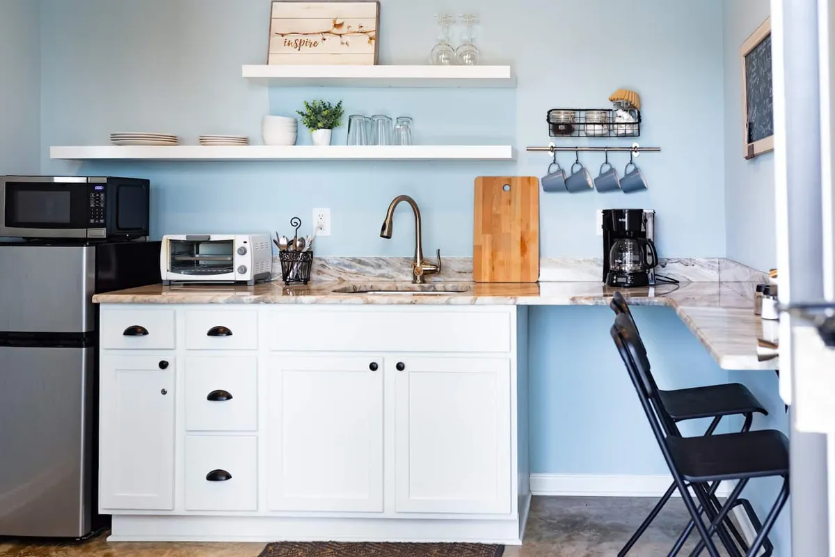 The image showcases a bright and tidy kitchen area featuring a sleek counter, modern sink, microwave, coffee maker, and open shelves adorned with tasteful decorations. The scene exudes a welcoming, cozy ambiance perfect for morning routines or casual meals, and hints at the apartment's carefully curated comforts.